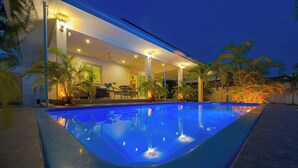 Piscina com área de estar coberta, palmeiras e um edifício com mesas ao ar livre.