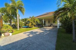 Uma casa com varanda coberta, entrada de automóveis em cascalho e palmeiras.