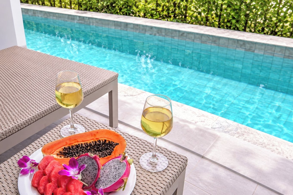 A poolside setting with a table, two glasses of white wine, a plate with sliced dragon fruit and watermelon, and a woven chair.