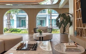 A modern lounge area with a chess table, comfortable seating, large windows, and a view of a building with balconies. - TITLE Serenity by TVC (Sakhu)