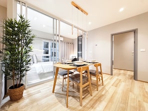 A modern dining area with a wooden table and chairs, a potted plant, and a view of the city through large windows. - TITLE Serenity by TVC (Sakhu)
