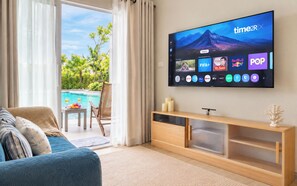 A modern living room with a large flat-screen TV mounted on the wall, a comfortable sofa, a wooden console table, and a view of a swimming pool. - TITLE Serenity by TVC (Sakhu)