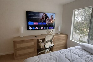 A hotel room with a bed, a desk with a chair, a TV mounted on the wall, and a window with a view of trees. - Newly Renovated Cozy bed and private bath with AC in beautiful San Diego (San Diego)