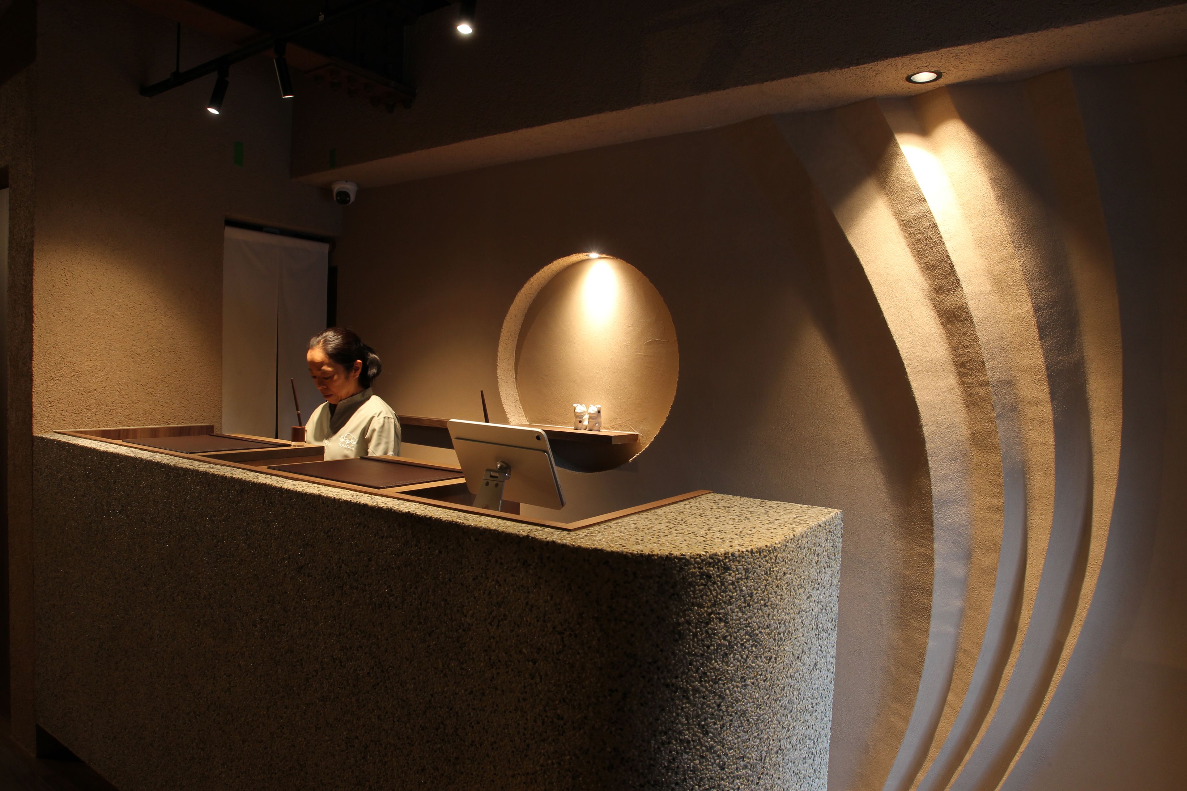 A reception desk with a person behind it, a round light fixture on the wall, and a textured wall design.