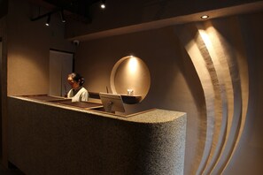 A reception desk with a person behind it, a round light fixture on the wall, and a textured wall design. - araijukuloop (Myoukou)