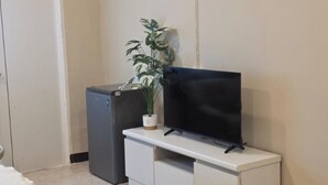 A bedroom with a bed, a TV on a white cabinet, a small refrigerator, and a potted plant. - Jomtien Beach Stay (Pattaya)