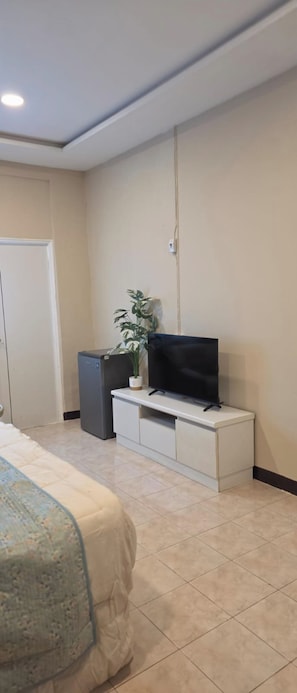 Un dormitorio con cama, televisor sobre un mueble blanco, refrigerador pequeño y una planta en maceta.