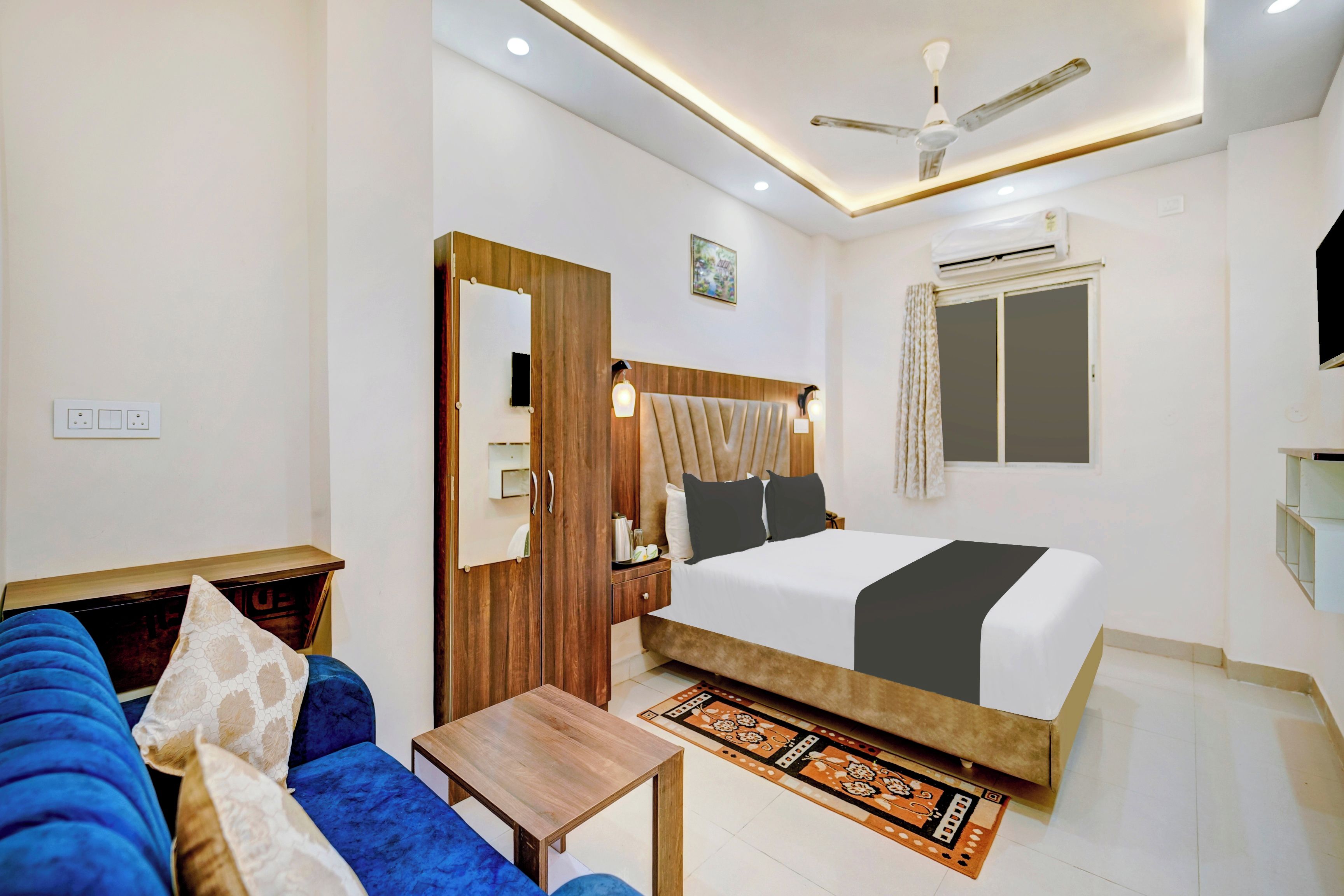 A hotel room with a bed, a sofa, a small table, a ceiling fan, an air conditioning unit, and a window with curtains.