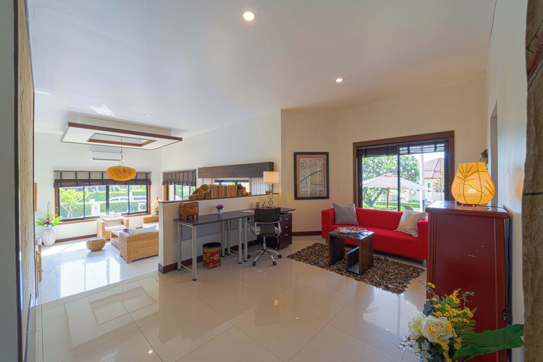 Amplia sala de estar con un sofá rojo, un escritorio y una mesa de comedor. Hay ventanas grandes que ofrecen luz natural y vistas al exterior.