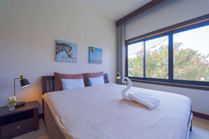 A hotel room with a large bed, a nightstand with a lamp, two wall art pieces, and a window with a view of pink flowers. - Stylish3BR PoolVilla,Prime Location-H146 (Hua Hin)