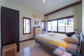 Quarto de hotel com cama, escrivaninha, abajur, cômoda e janela com persianas.