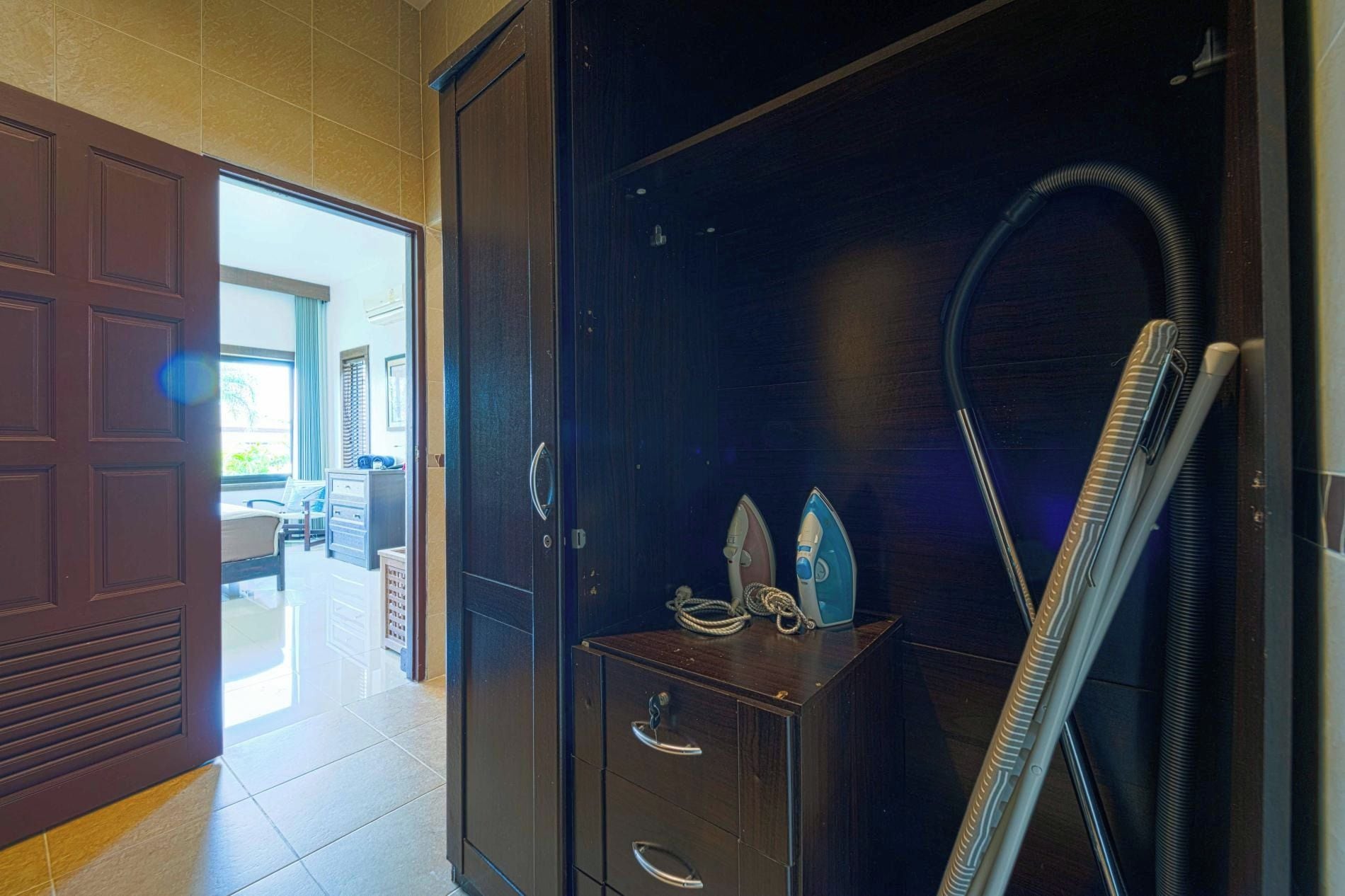 Quarto de hotel com guarda-roupa de madeira escura, uma secretária pequena com gavetas e um aspirador de pó.