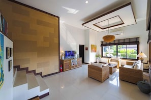 A spacious living area with a high ceiling, a television, wicker furniture, and a large window offering a view of the outdoors. - Stylish3BR PoolVilla,Prime Location-H146 (Hua Hin)