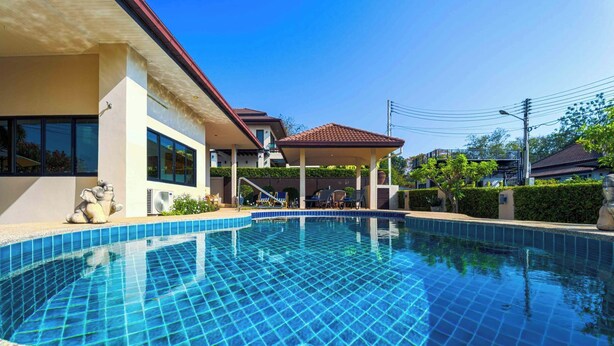Une piscine avec un bord carrelé, un patio couvert et une maison dotée de grandes fenêtres et d’un toit rouge.