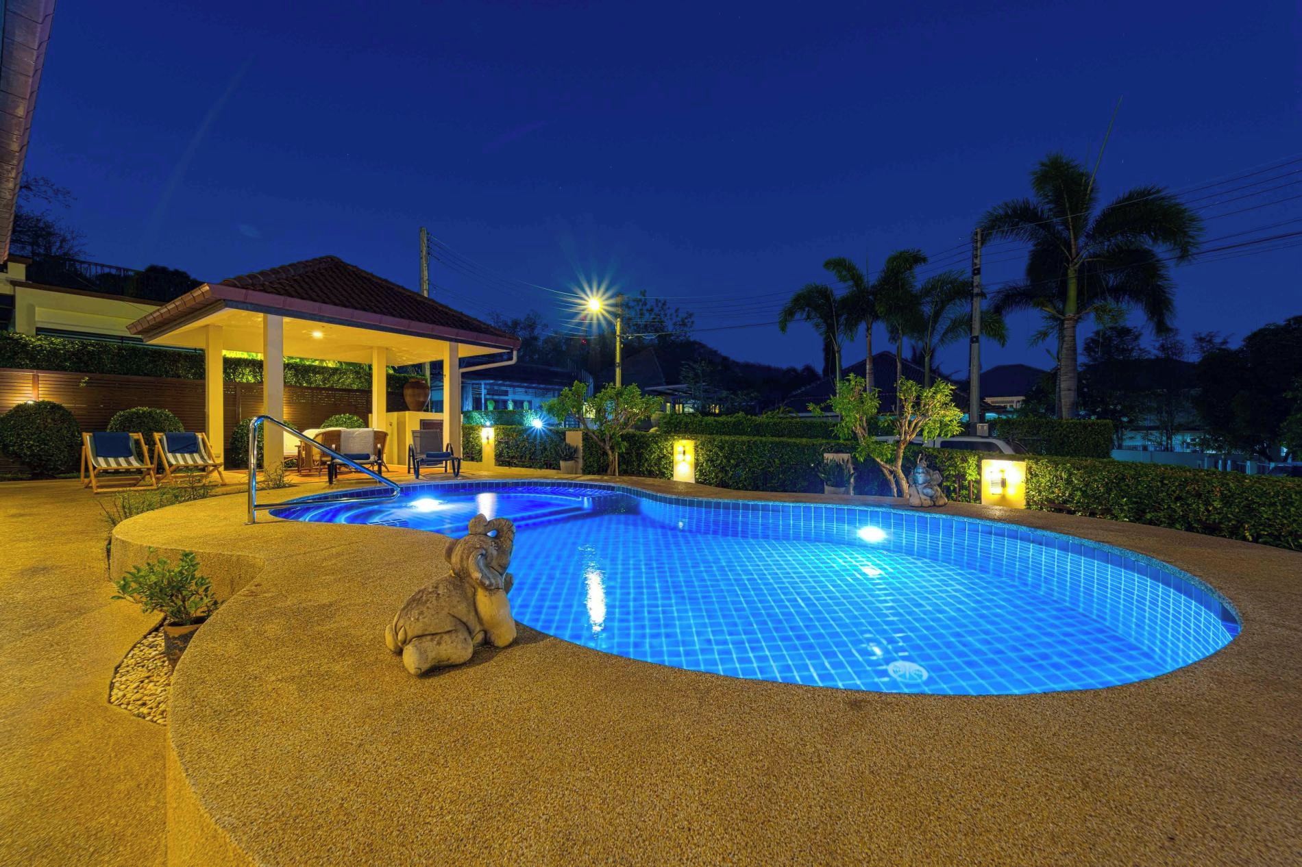 Piscina com área central rebaixada, rodeada por um espaço coberto com cadeiras e uma mesa, e um pequeno edifício com telhado.