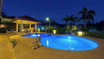 Piscina com área central rebaixada, rodeada por um espaço coberto com cadeiras e uma mesa, e um pequeno edifício com telhado.