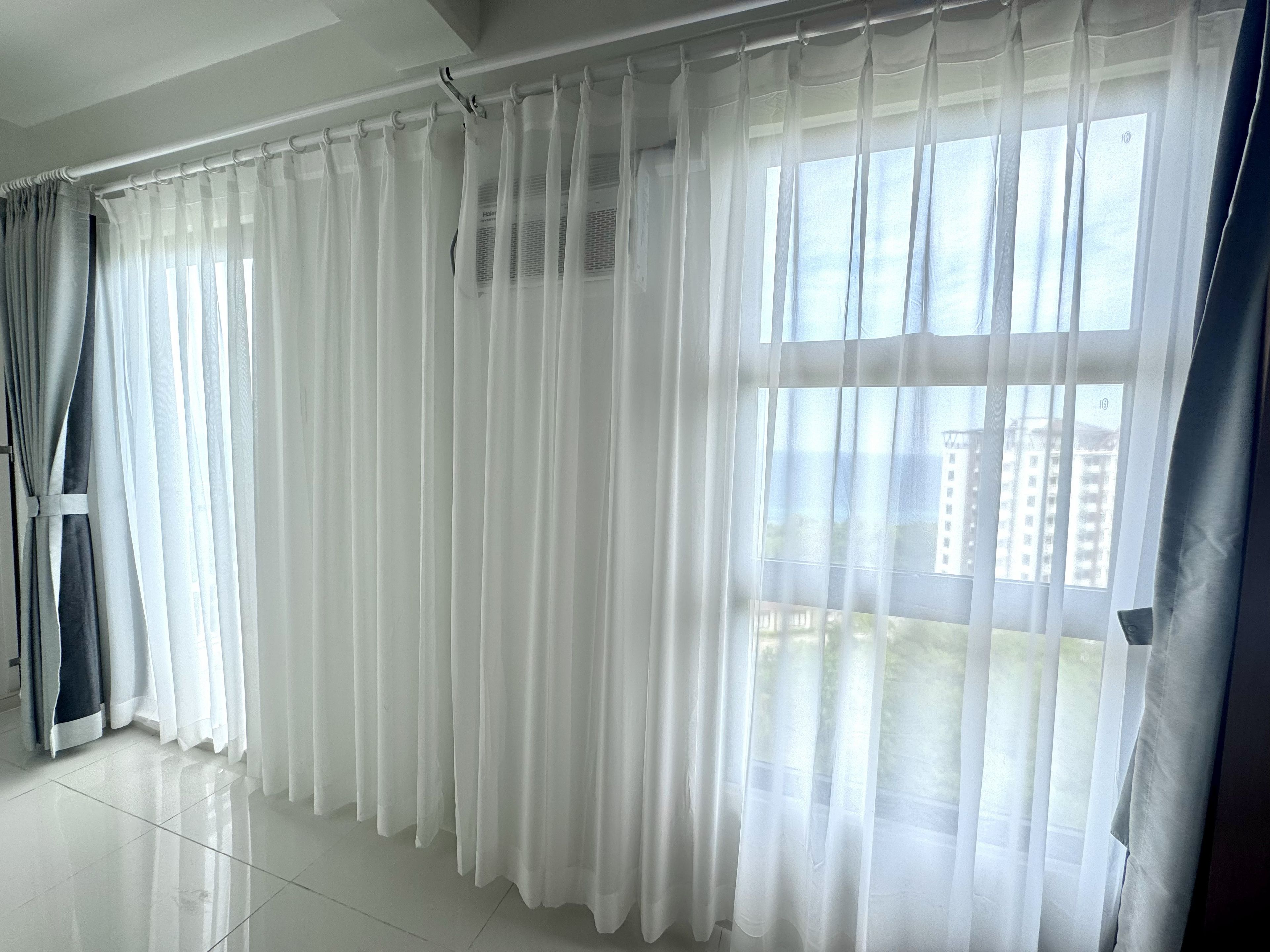 Ein Zimmer mit einem großen Fenster, das mit transparenten weißen Vorhängen drapiert ist, einem gefliesten Boden und Blick auf die Stadt.
