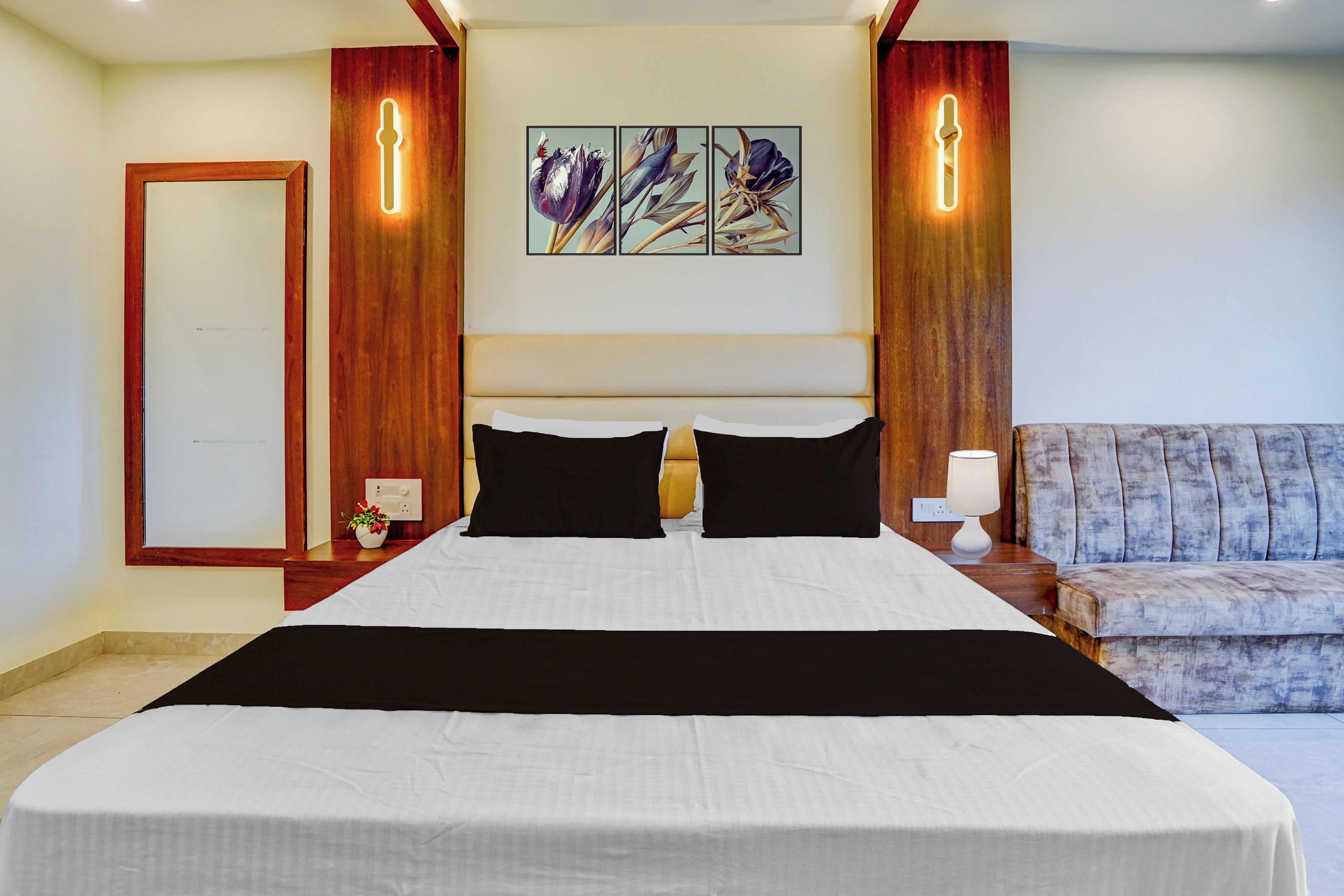Habitación de hotel con una cama grande, cabecero de madera, mesita de noche con lámpara, zona de estar con un banco acolchado y una obra de arte colgada en la pared.