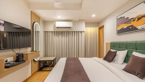 Quarto de hotel com cama, escrivaninha, cadeira, televisão, aparelho de ar condicionado e um quadro na parede.