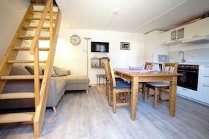 A cozy living space with a wooden staircase, a sofa, a dining table with chairs, a kitchen area, and a television on the wall. - Fischerhus Krummin - Thatched-roof house with a private sauna (Krummin)