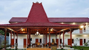 Um edifício tradicional com telhado de barro avermelhado, varanda coberta com pilares de madeira e área de jardim com plantas em vasos.