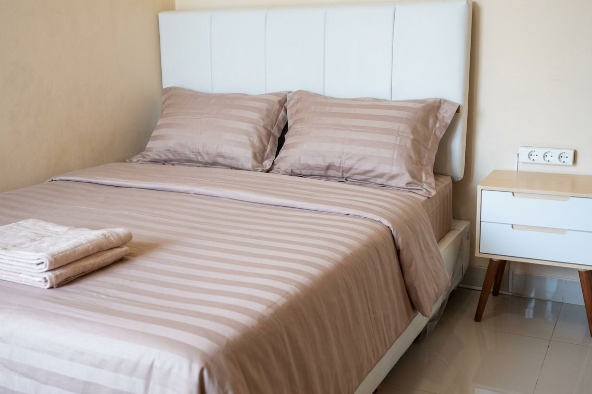 A neatly made bed with striped bedding and pillows, a bedside table with a lamp, and a power outlet on the wall.