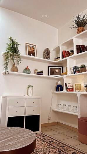 Ein Zimmer mit weißen Wänden, einem weißen Schrank mit Schubladen, einem runden Tisch und Regalen mit Büchern und Dekoartikeln.
