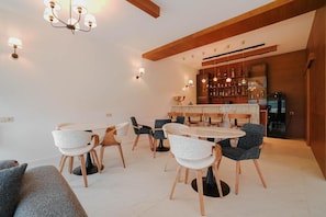 Un comedor moderno con mesas y sillas redondas, una zona de bar con estantes llenos de botellas, y una cocina al fondo.