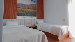 A hotel room with two beds, wooden flooring, a large window with curtains, and a wall-mounted light fixture. - Mikheilis Marani Boutique Hotel  (Kvareli)
