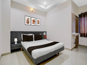 A hotel room with a bed, nightstand, lamp, wall-mounted artwork, and a bathroom with a sink and shower curtain. - Deluxe Hotel Sai Dhanlaxmi (Rahata)