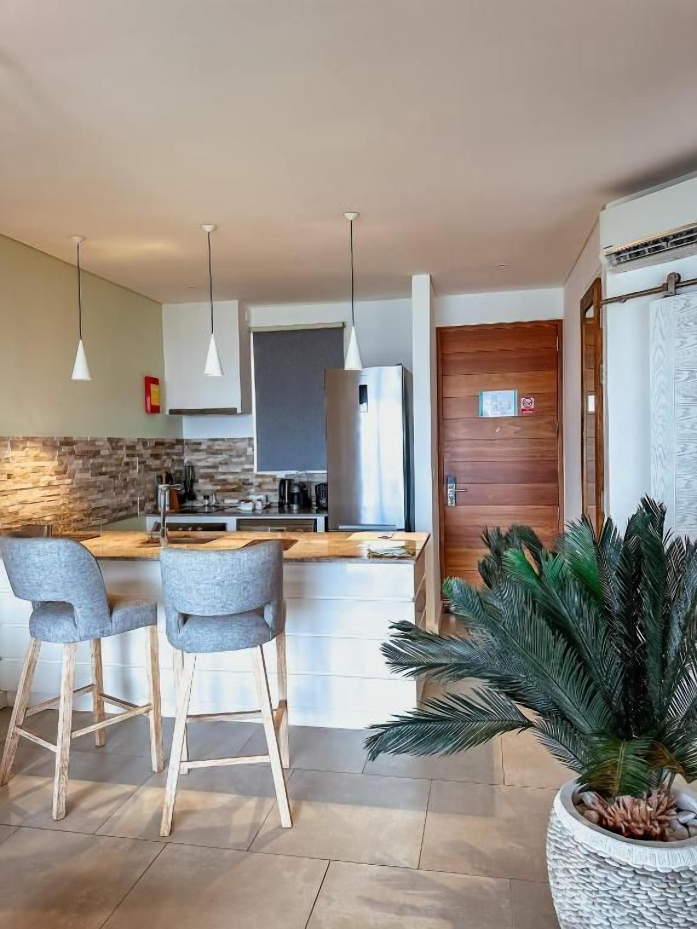 Una cocina moderna con una isla central, taburetes de bar, iluminación colgante y una planta grande en maceta.