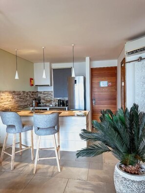 Una cocina moderna con una isla central, taburetes de bar, iluminación colgante y una planta grande en maceta.
