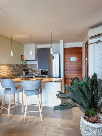 A modern kitchen with a central island, bar stools, pendant lighting, and a large potted plant.
