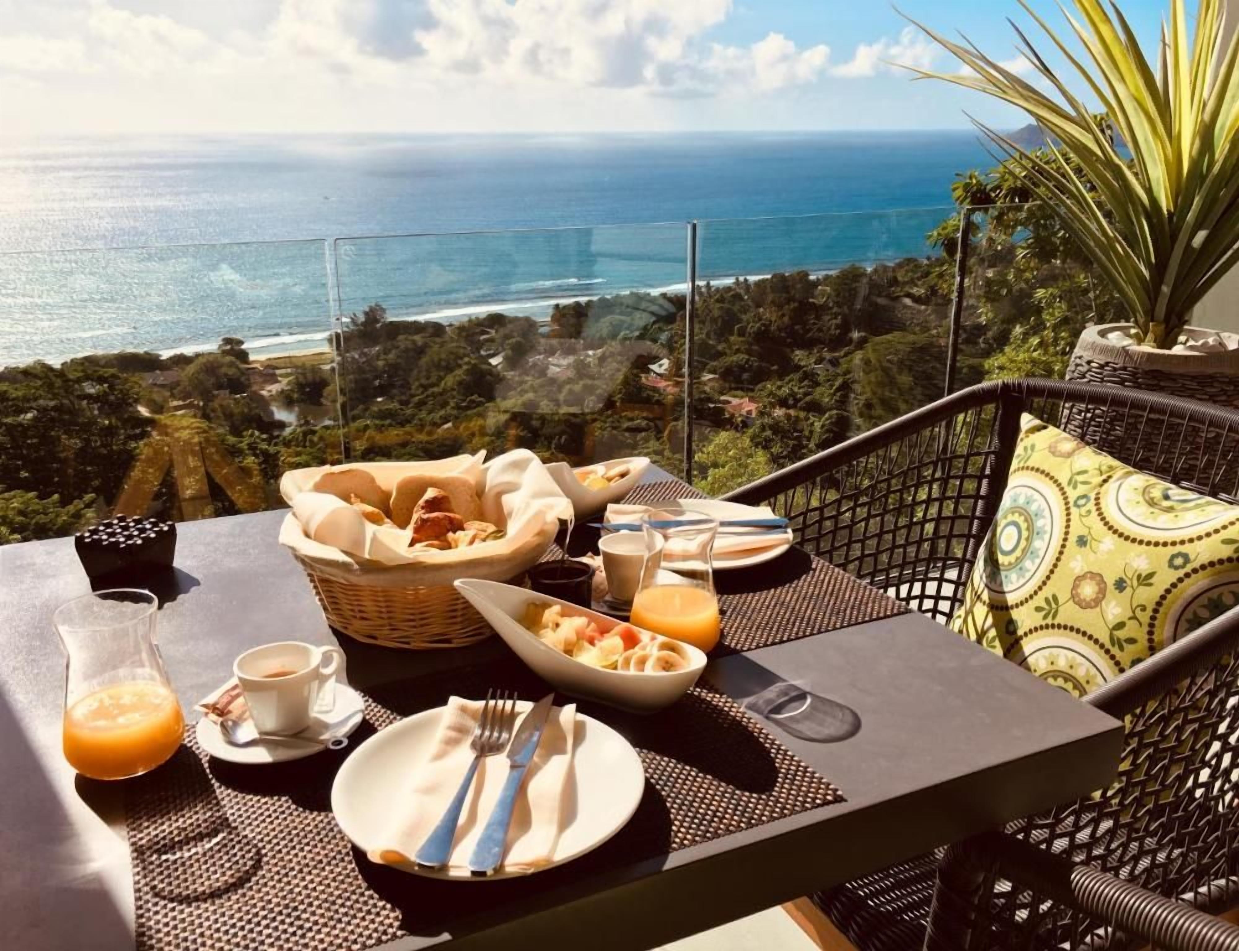 Una mesa lista para el desayuno con vista al océano, una cesta de pan, un tazón de pasta y un vaso de jugo de naranja.