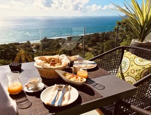 Una mesa lista para el desayuno con vista al océano, una cesta de pan, un tazón de pasta y un vaso de jugo de naranja.