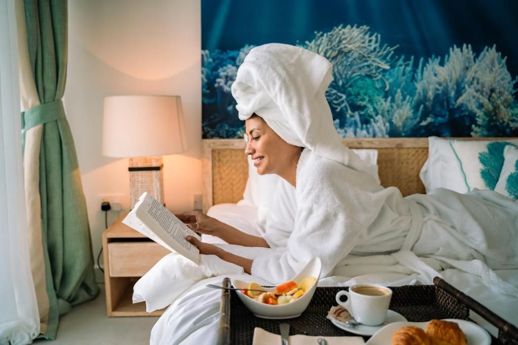 Eine Person in einem weißen Bademantel liest ein Buch in einem Hotelzimmer, das ein Bett, einen Nachttisch, eine Lampe, eine Obstschale und eine Kaffeetasse beinhaltet.