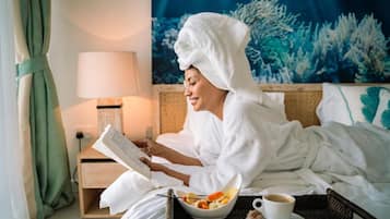 Eine Person in einem weißen Bademantel liest ein Buch in einem Hotelzimmer, das ein Bett, einen Nachttisch, eine Lampe, eine Obstschale und eine Kaffeetasse beinhaltet.