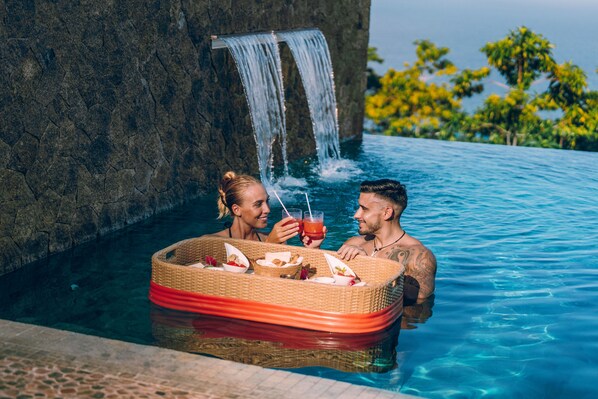 A couple relaxing in a pool with a waterfall feature, surrounded by lush greenery.