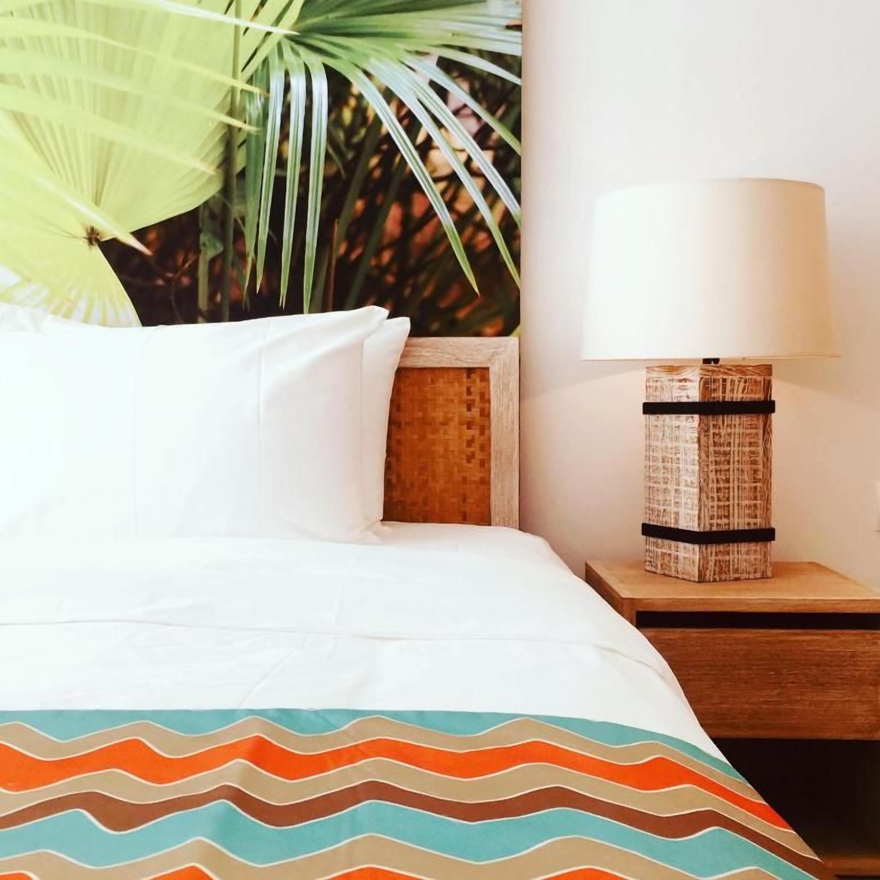 Una cama con ropa blanca, un plaid estampado, una mesita de noche con lámpara y una pared con una planta.