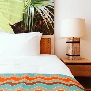 Una cama con ropa blanca, un plaid estampado, una mesita de noche con lámpara y una pared con una planta.