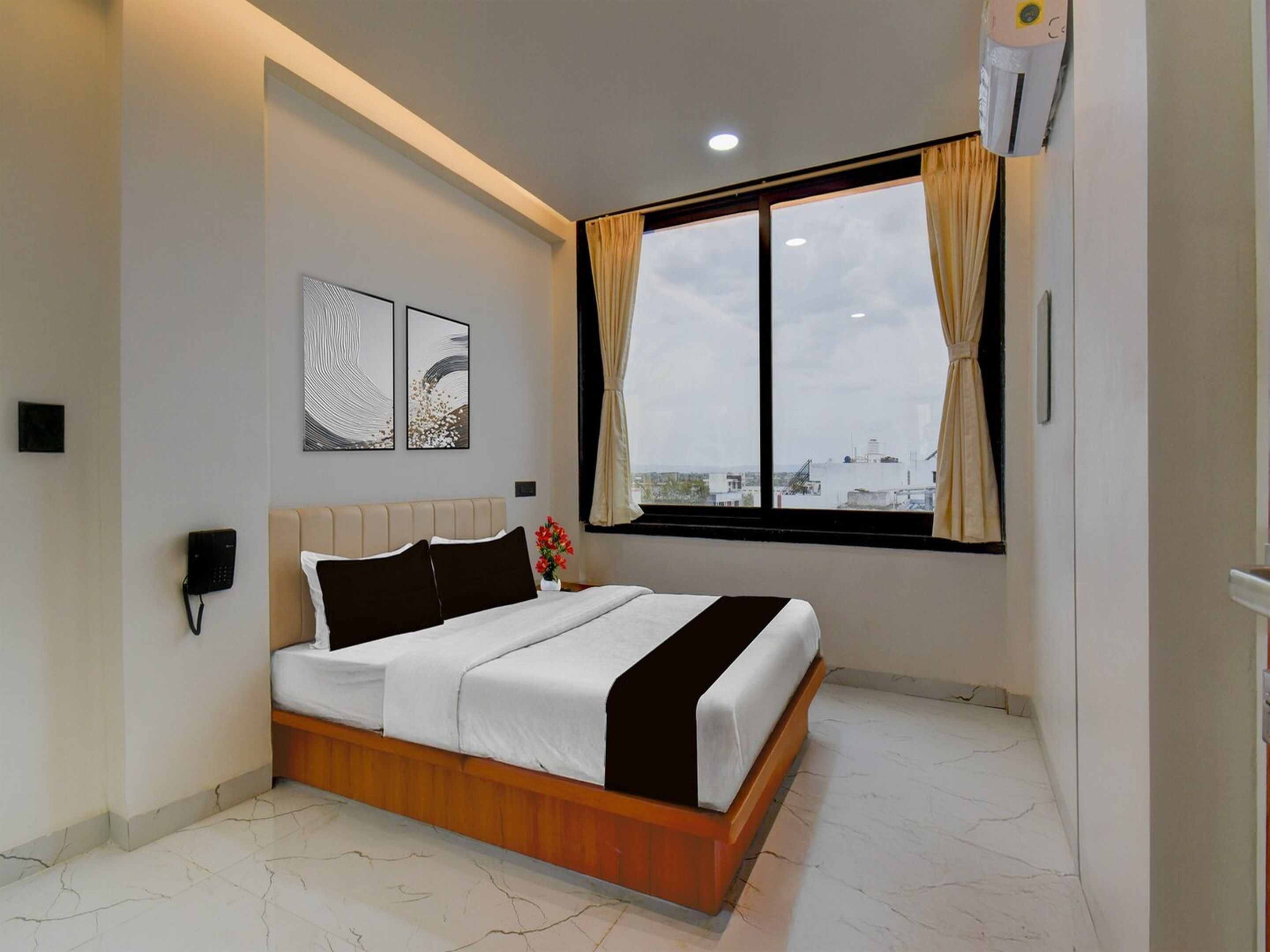 A modern bedroom with a bed, a phone, artwork, a window with curtains, and a city view.
