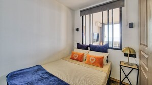 A bedroom with a bed, a window with blinds, a bedside lamp, and a view of the outdoors. - "Horizon Bleu" One-bedroom apartment with balcony, ocean view, pool, and parking (Cap-d'Ail)