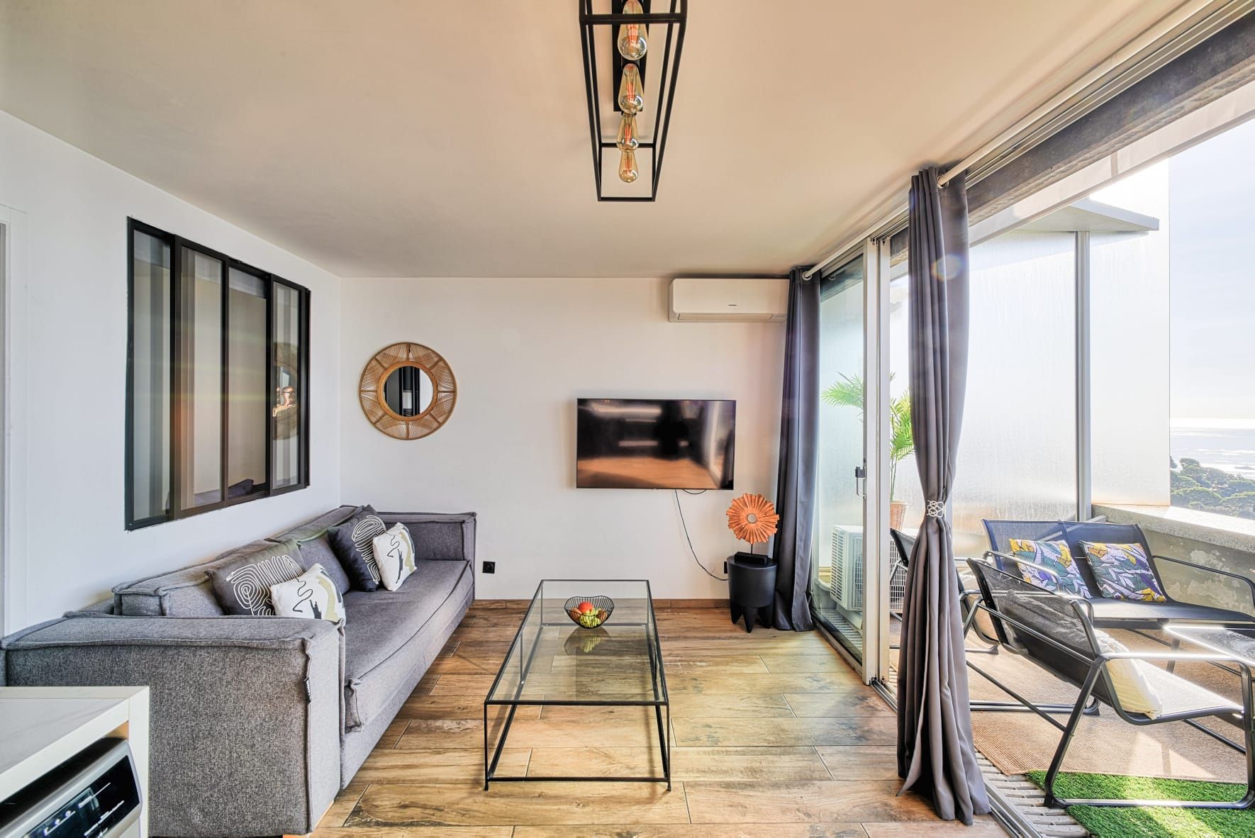 A modern living room with a sofa, a coffee table, a TV, and a balcony with outdoor seating.