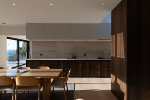A modern kitchen with a large island, wooden chairs, and a view of the outdoors. - Porto Sanctuary | Tranquil Coastal Luxury with Sweeping Views (Nelson)