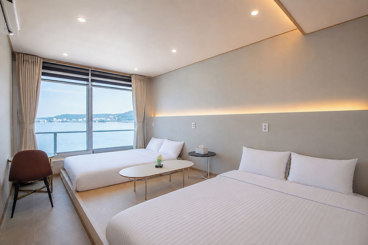A hotel room with two beds, a small table, a chair, and a view of the sea.