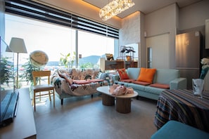 A modern living room with a sofa, armchairs, a coffee table, and a dining table. There is a large window with blinds, a lamp, and a globe on a stand. - OCEANSUITE (Busan)