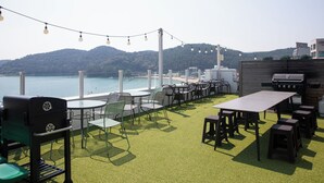 An outdoor seating area with tables and chairs, a grill, and a view of the sea and hills. - OCEANSUITE (Busan)