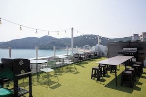 An outdoor seating area with tables and chairs, a grill, and a view of the sea and hills. - OCEANSUITE (Busan)