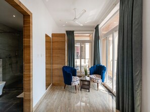 Ein modernes Hotelzimmer mit Dusche, einer Holzwand, einem Deckenventilator, einer Sitzecke mit zwei Stühlen und einem kleinen Tisch sowie großen Fenstern mit Vorhängen.
