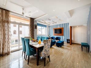 Ein modernes Hotelzimmer mit Essbereich, einer Sitzecke mit blauen Polsterstühlen, einem an der Wand befestigten Flachbildfernseher und einem großen Fenster mit Vorhängen.